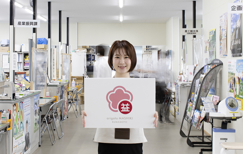 被災地から感謝を伝える取り組み「arigato MASHIKI」（2021年度グッドデザイン賞受賞）のイメージ写真。熊本県益城町のコミュニケーションマークを持つ町役場職員。熊本県益城町役場・仮設庁舎にて撮影。
